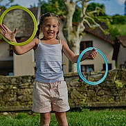 Lola a rejoint le concours — aidez-le/la à gagner de superbes lots ! child, girl, smiling, outdoor, grass, hoops, playing, summer, casual_clothing, shorts, tank_top, happy, daylight, trees, stone_wall, house, nature, recreation, fun, leisure