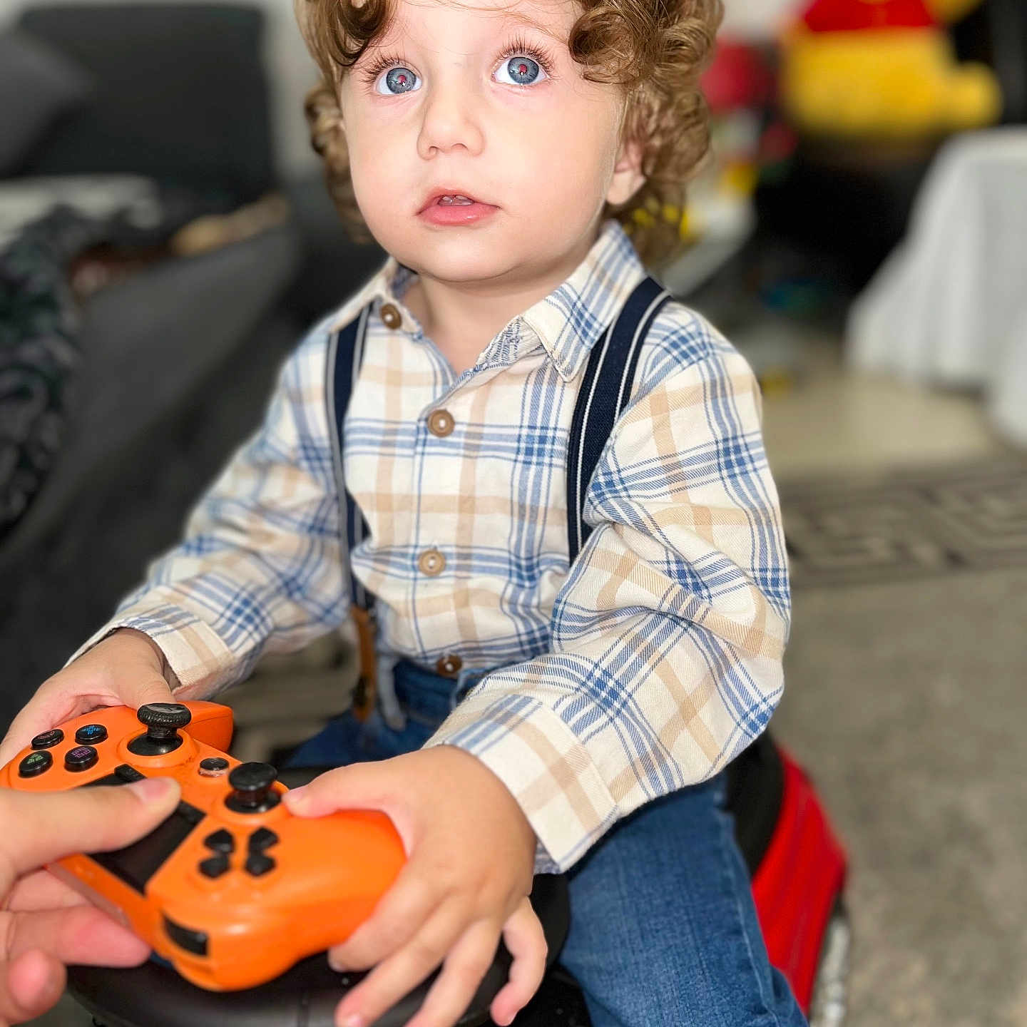 Elys Dragulin a rejoint le concours — aidez-le/la à gagner de superbes lots ! blue_eyes, child, curly_hair, face, focused, game_controller, hands, indoor, jeans, living_room, orange, person, plaid_shirt, playing, plush_toy, red, seated, suspenders, toddler, toy_car