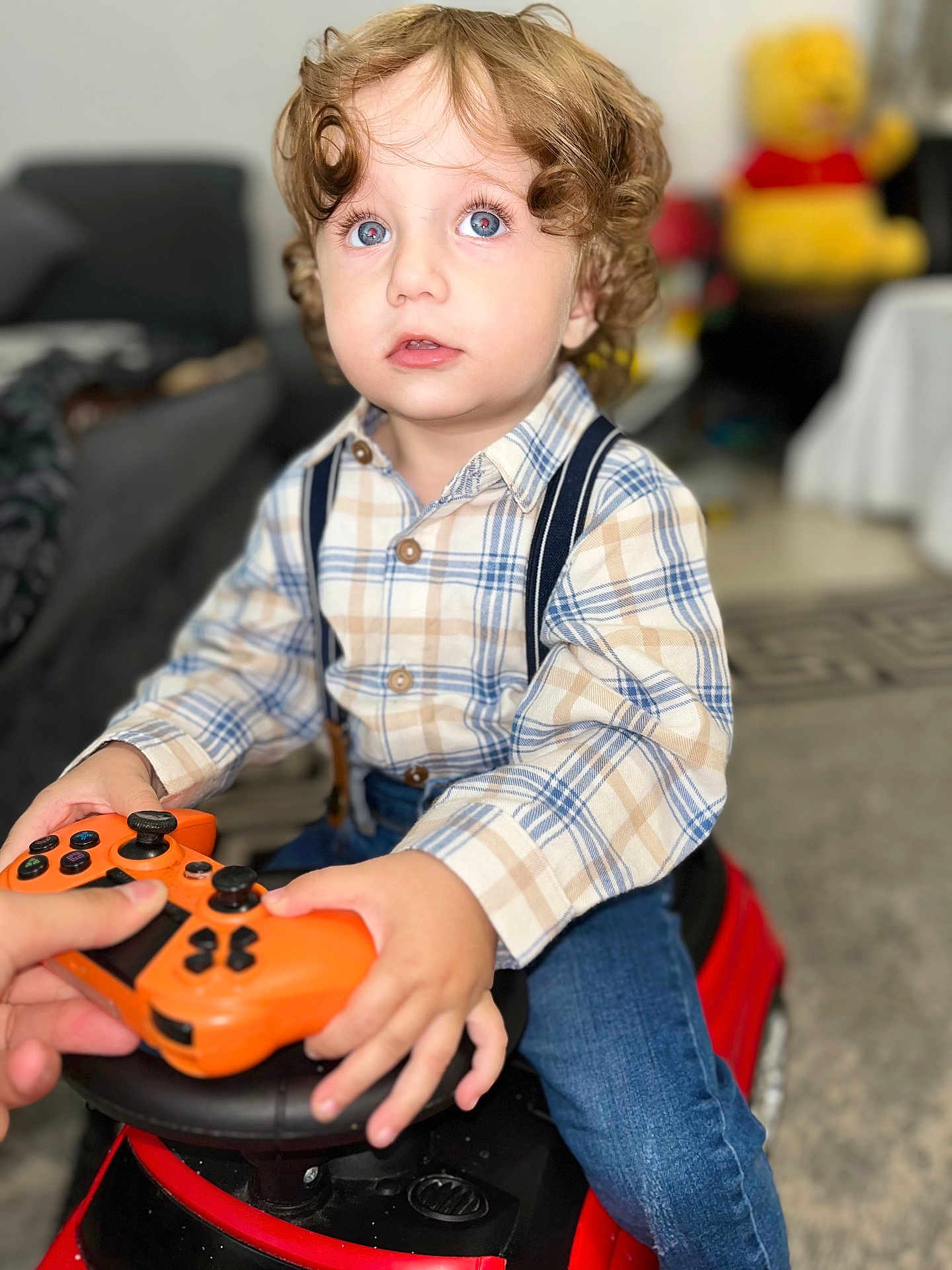 Elys Dragulin a rejoint le concours — aidez-le/la à gagner de superbes lots ! blue_eyes, child, curly_hair, face, focused, game_controller, hands, indoor, jeans, living_room, orange, person, plaid_shirt, playing, plush_toy, red, seated, suspenders, toddler, toy_car