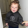 black_clothing, bright_eyes, casual_clothing, child, curly_hair, door, face_paint, floor, hand_holding, handprints, happy, hoodie, indoor, person, portrait, smiling, standing, toddler, wall, young_child