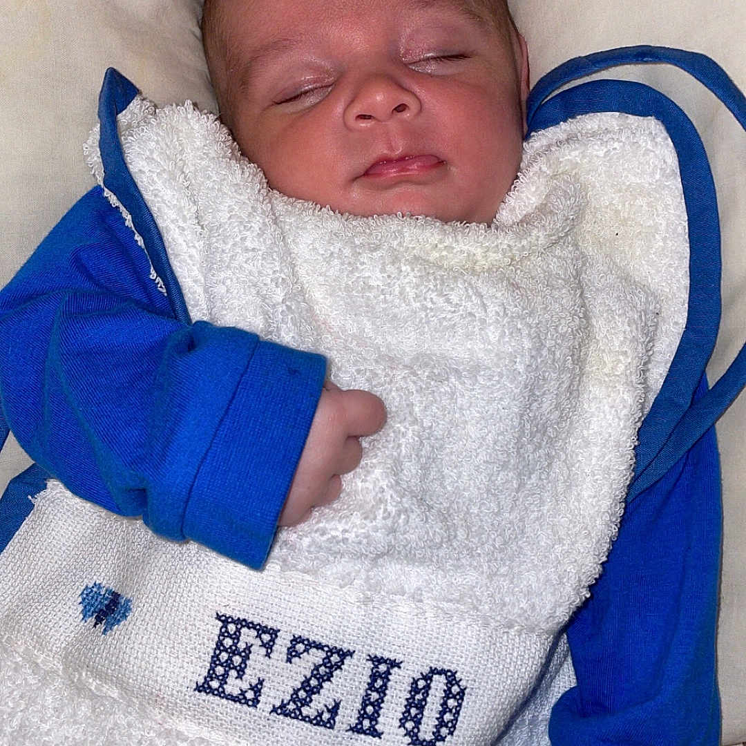 Ezio participe au concours pour gagner de l'argent avec cette photo : baby, bib, blue_clothing, child, closeup, cute, embroidered_text, face, floral_background, hand, indoors, infant, newborn, peaceful, pillow, portrait, resting, sleep, sleeping, soft_texture