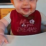 Blakely is registered to the contest to win money with this photo: baby, bib, child, clothing, cute, expression, eyes, face, food, happy, high_chair, indoor, infant, kitchen, messy, mouth, person, smiling, table, tongue
