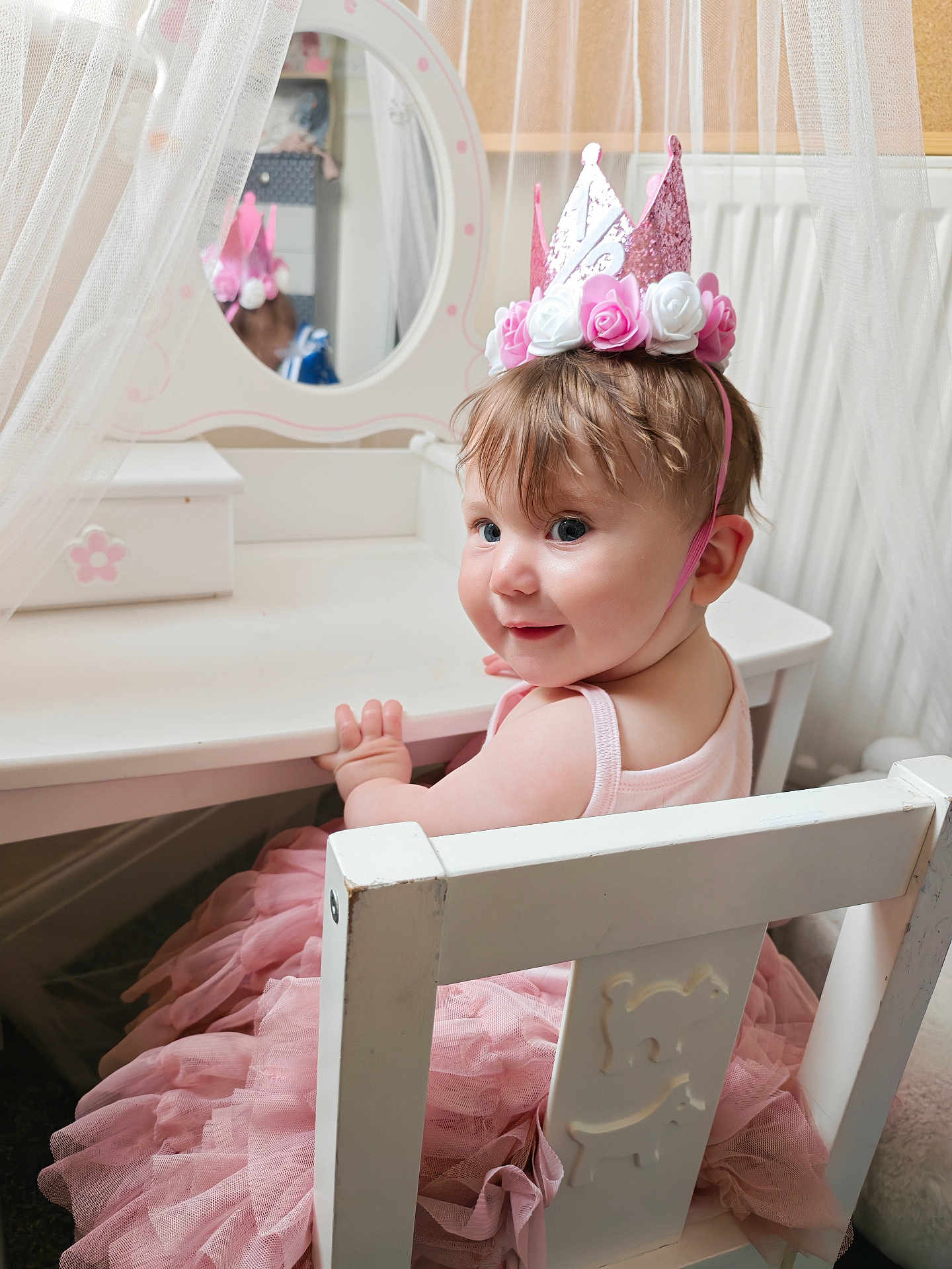 Isabella is registered to the contest to win money with this photo: toddler, child, pink_crown, tutu_dress, white_chair, vanity_mirror, curtains, smile, indoor, furniture, face, person, cute, birthday_hat, playroom, pink, dress, hair, baby, happy
