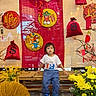 child, bench, flowers, yellow_flowers, red_decorations, money_bags, bamboo, t_shirt, blue_pants, sneakers, indoor, traditional_art, festive, plant, floor, person, portrait, young_child, cultural, bench_seating