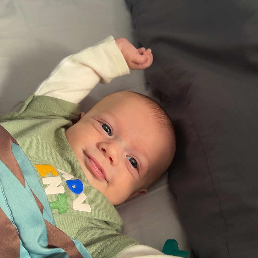 Daxton is registered to the contest to win money with this photo: baby, bed, bedding, blanket, close_up, clothing, cozy, cute, eyes, face, hand, infant, lying_down, nursery_item, onesie, pacifier, pillow, portrait, skin, smiling