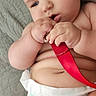 baby, infant, diaper, red_strap, hand, skin, face, curious, lying_down, soft_surface, chubby, cute, child, closeup, indoors, expression, chewing, rolls, person, young