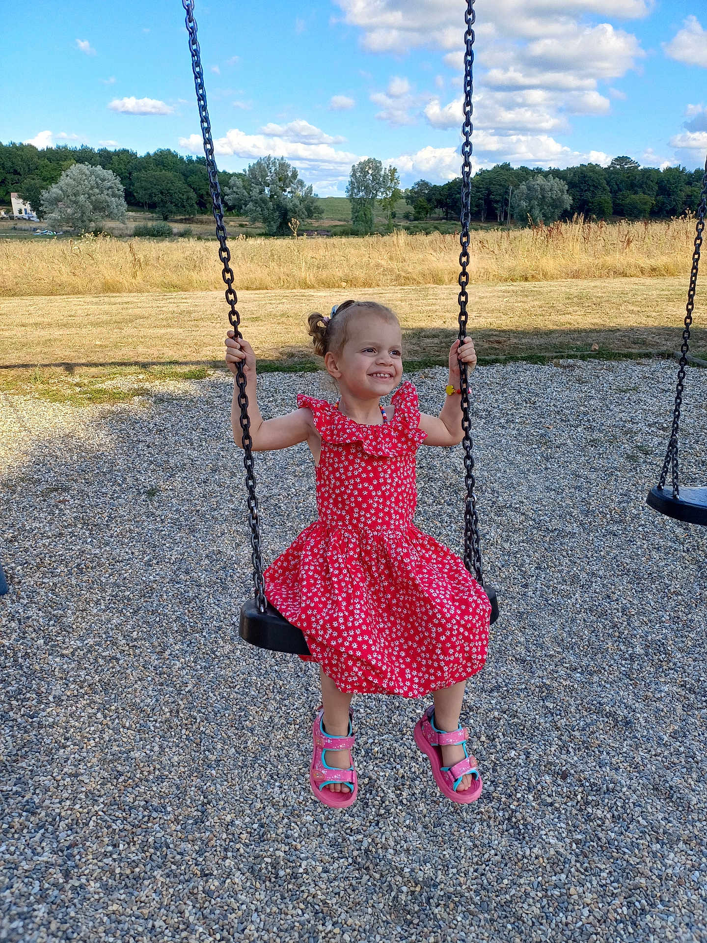 Lou-ann participe au concours pour gagner de l'argent avec cette photo : child, girl, swing, playground, red_dress, smiling, sandals, gravel, outdoor, daylight, blue_sky, clouds, trees, grass, field, happy, summer, nature, person, recreation