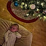 baby, blanket, celebration, child, christmas_tree, cushion, decorations, festive, floor, holiday, home, indoors, infant, lights, ornaments, pajamas, person, relaxing, sleepwear, wooden_floor