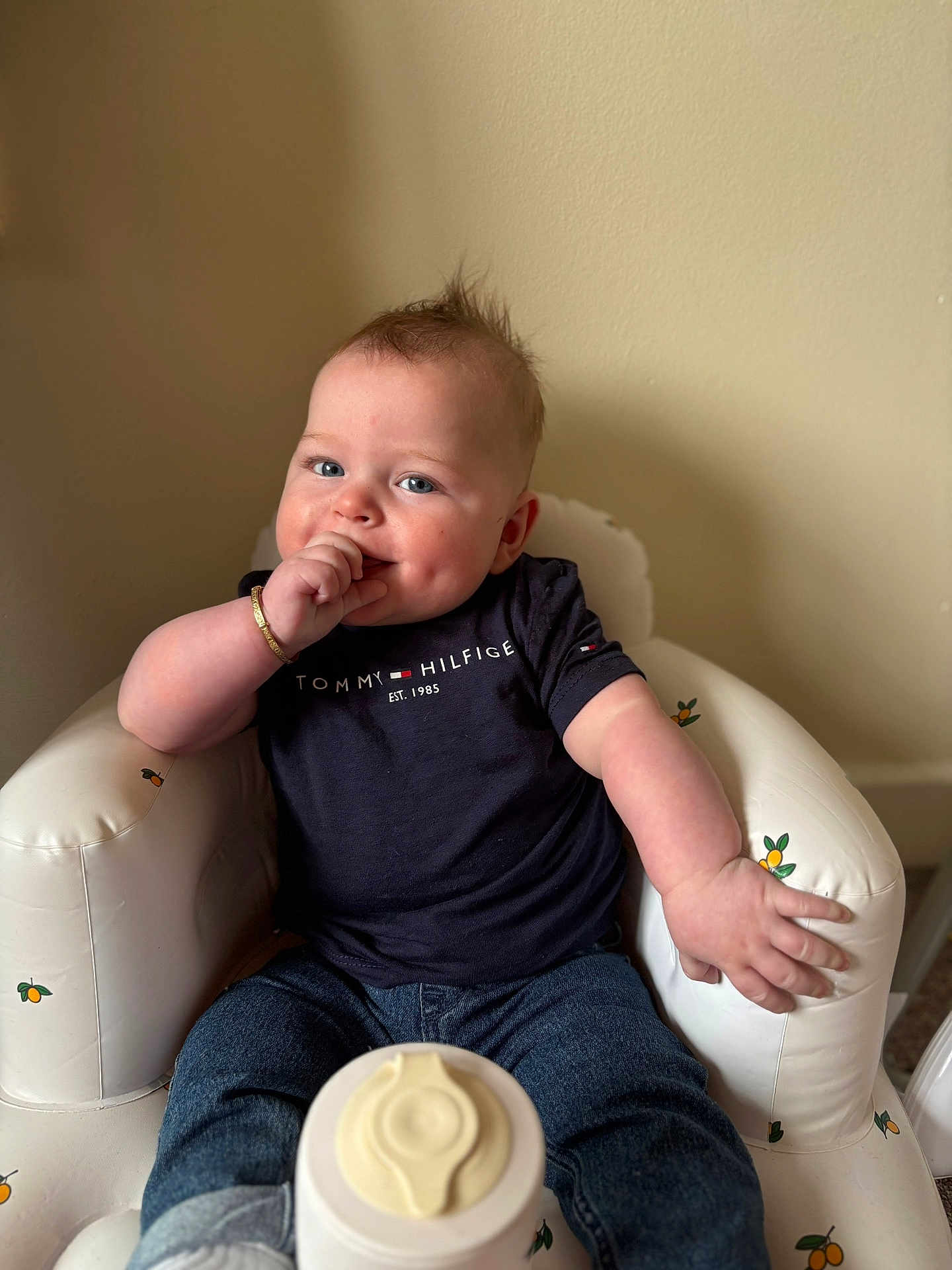 Ralphie joined the competition — help win amazing prizes! baby, infant, child, sitting, thumb_sucking, navy_shirt, jeans, white_chair, orange_pattern, curly_hair, bracelet, indoor, cute, smiling, portrait, young_child, casual_clothing, happy, plush_chair, relaxed