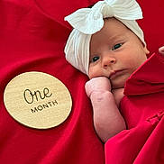 Eden is registered to the contest to win money with this photo: baby, infant, one_month, headband, white_bow, red_blanket, red_background, wooden_plaque, hand, face, portrait, newborn, cute, lying_down, skin, expression, child, closeup, soft, resting