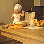 apron, baby, baking, bowl, bread, chef_hat, child, cooking, counter, eggs, flour, food, happy, indoor, kitchen, messy, person, playful, smile, wooden_table