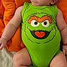 baby, infant, onesie, green_clothing, cartoon_face, orange_cushion, baby_socks, smiling, lying_down, cute, child, indoor, playful, happy, young_child, red_bracelet, skin, head, face, expression