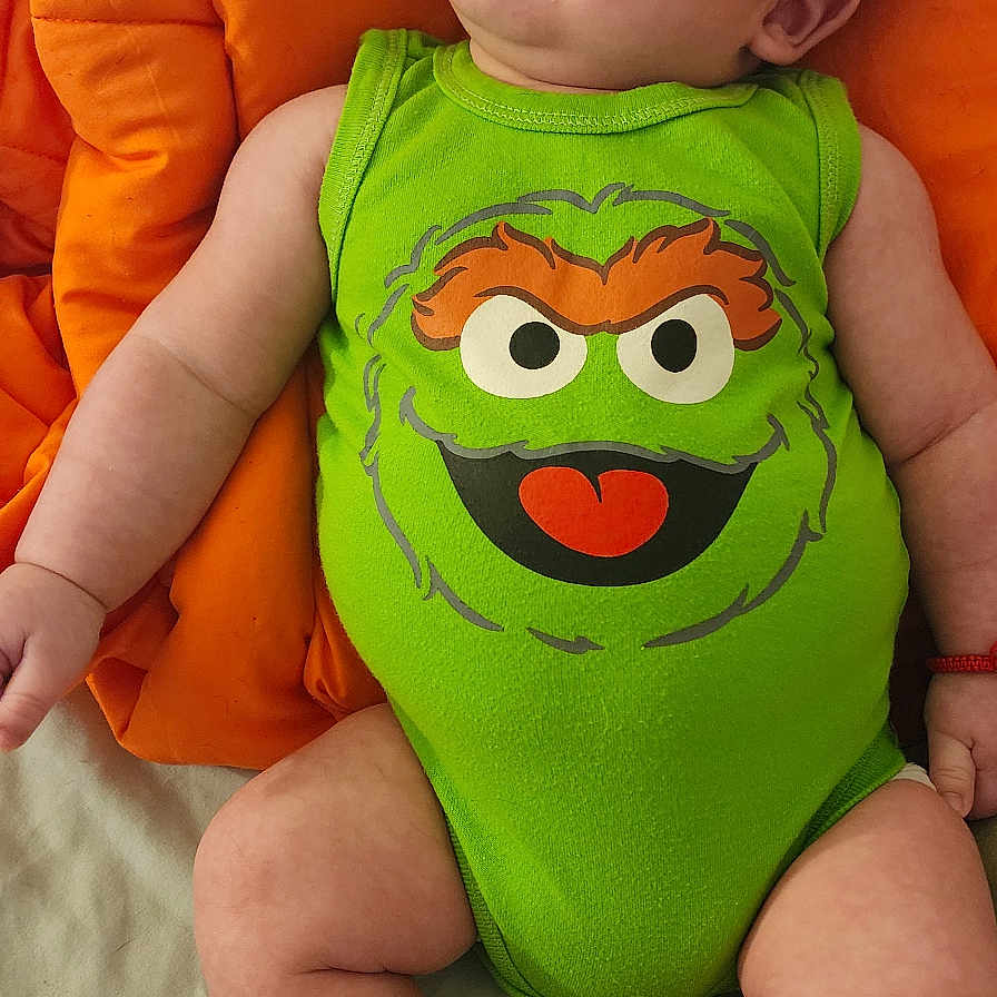 Idenshi joined the competition — help win amazing prizes! baby, infant, onesie, green_clothing, cartoon_face, orange_cushion, baby_socks, smiling, lying_down, cute, child, indoor, playful, happy, young_child, red_bracelet, skin, head, face, expression