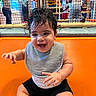León is registered to the contest to win money with this photo: baby, child, smiling, curly_hair, bib, black_shorts, orange_surface, indoor_playground, net, play_area, happy, sitting, person, infant, casual_clothing, bracelet, background_children, bright_colors, cute, fun