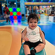 León joined the competition — help win amazing prizes! toddler, child, smiling, curly_hair, white_shirt, black_shorts, socks, chair, indoor, play_area, colorful_lights, blurred_background, happy, person, portrait, seated, fun, casual_clothing, toy_area, bright_colors
