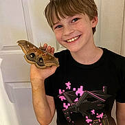 Brandon is registered to the contest to win money with this photo: child, boy, smile, moth, insect, hand, tshirt, indoor, door, short_hair, freckles, happy, portrait, animal, animal_in_hand, casual_clothing, young, light_skin, standing, nature