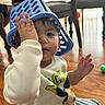 toddler, child, indoor, floor, plastic_basket, hat, hand, face, sweater, cartoon_character, wooden_floor, chair, window, playful, cute, young_child, clothing, person, expression, funny