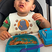 Noah joined the competition — help win amazing prizes! toddler, child, high_chair, bib, cookie_monster, food, spaghetti, plate, spoon, toy_truck, expression, messy, indoor, table, green_shirt, face, hair, hand, person, meal
