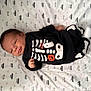 baby, infant, smile, clothing, skeleton_outfit, halloween, blanket, pattern, car_pattern, lying_down, cute, young_child, face, head, hand, footwear, sock, black_clothing, white_pattern, indoors