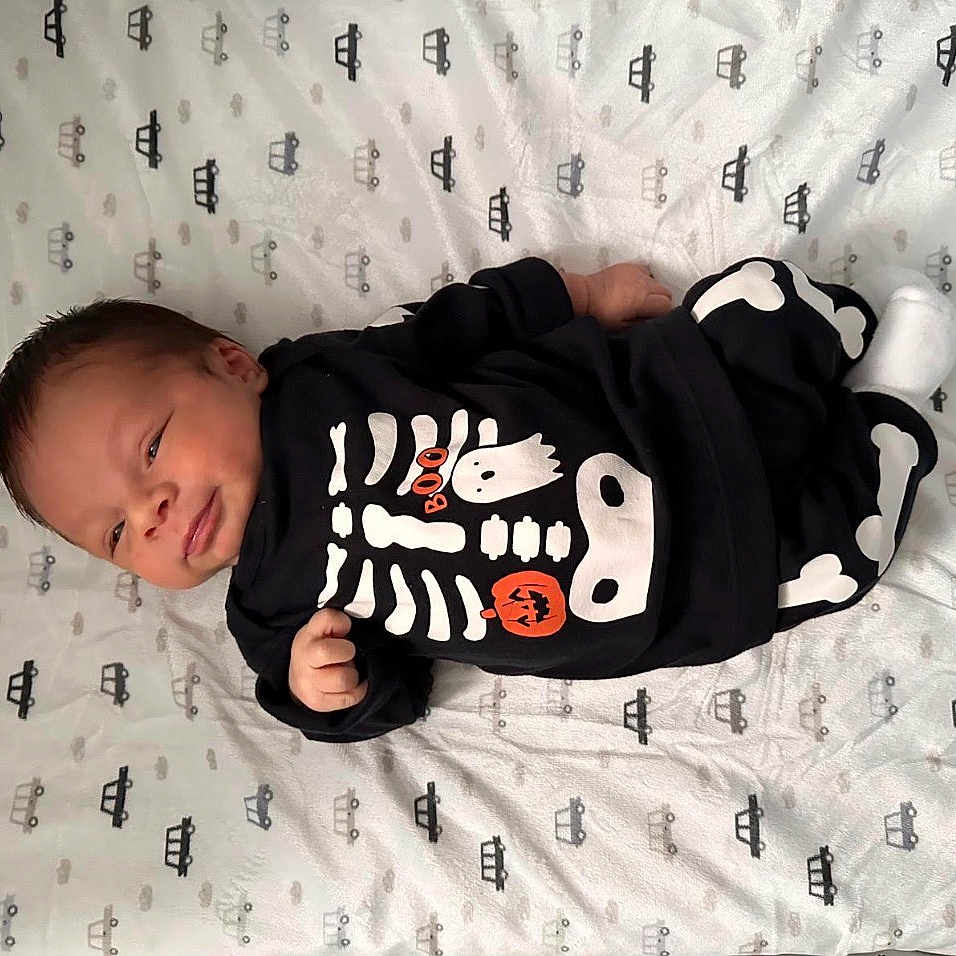 Maximiliano is registered to the contest to win money with this photo: baby, black_clothing, blanket, car_pattern, clothing, cute, face, footwear, halloween, hand, head, indoors, infant, lying_down, pattern, skeleton_outfit, smile, sock, white_pattern, young_child