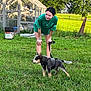 girl, puppy, dog, green_shirt, leash, grass, outdoor, greenhouse, garden, sunlight, smile, young_person, pet, nature, summer, casual_clothing, trees, playful, animal, field