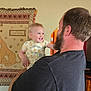 Micha is registered to the contest to win money with this photo: baby, beard, blue_eyes, child, clothing, cute, happy, holding, home, indoor, interaction, joyful, lion_king, man, person, portrait, short_sleeve, smile, tapestry, wall_hanging