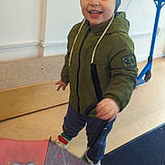 Mylo is registered to the contest to win money with this photo: child, smiling, umbrella, hat, indoor, jacket, scooter, floor, bench, toy, happy, boy, clothing, playful, person, footwear, headwear, colorful, face, standing