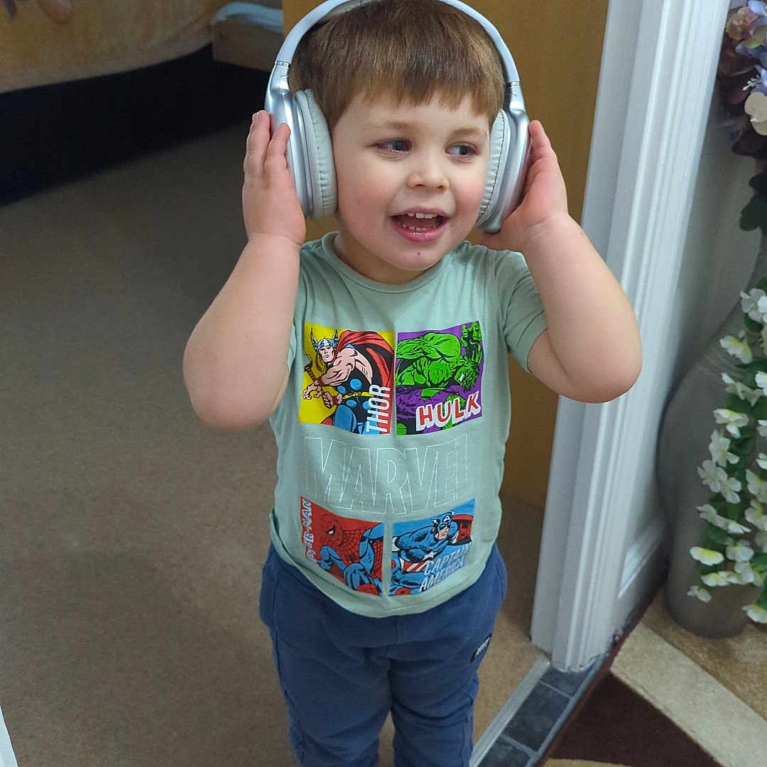 Mylo is registered to the contest to win money with this photo: bed, boy, carpet, carpet_rug, child, decor, doorway, flower_vase, headphones, home, indoor, marvel, pants, playful, short_hair, smiling, socks, standing, striped_socks, tshirt