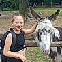 child, girl, donkey, animal, petting, farm, outdoor, tree, fence, grass, black_clothing, smiling, backpack, ears, nature, daylight, cute, young, friendly, rural