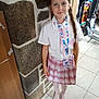 child, girl, braid, school_uniform, plaid_skirt, white_shirt, lanyard, indoor, stone_wall, tiled_floor, socks, toy_scooter, smiling, standing, person, young, casual, portrait, home, accessory