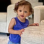 blond_hair, blue_eyes, child, curious, curly_hair, cute, expression, furniture, hairclip, hand, home, indoor, living_room, ottoman, portrait, purple_top, skin, standing, star_shorts, toddler
