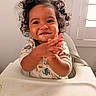 toddler, child, curly_hair, high_chair, messy_face, clapping_hands, smiling, indoor, baby_food, cute, happy, person, infant, apparel, long_sleeves, light_switch, window, face, skin, hands
