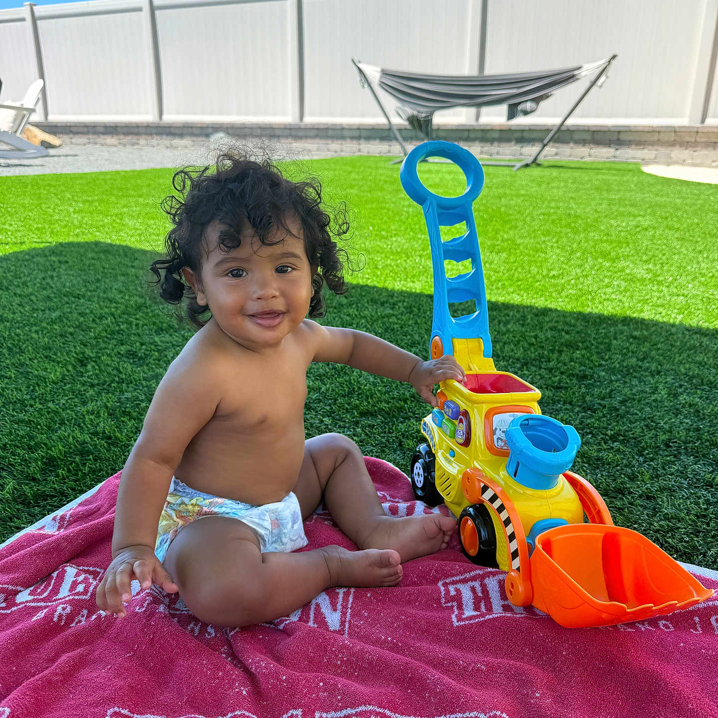 Julian joined the competition — help win amazing prizes! toddler, child, toy, excavator, red_towel, grass, outdoor, sunny, blue_sky, curly_hair, smiling, bare_chest, diaper, playtime, fence, backyard, plastic_toy, happy, sitting, daytime