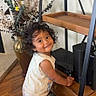 Julian is registered to the contest to win money with this photo: toddler, child, curly_hair, smiling, barefoot, wooden_floor, shelf, electronic_device, vase, dried_flowers, balloon, indoor, person, furniture, happy, standing, room, decor, baby, playful