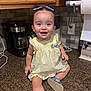 toddler, child, baby, kitchen, countertop, yellow_dress, blue_bows, blue_sandals, smiling, sitting, coffee_maker, paper_towels, cabinet, tile_wall, cute, happy, indoor, person, hair_bows, playful