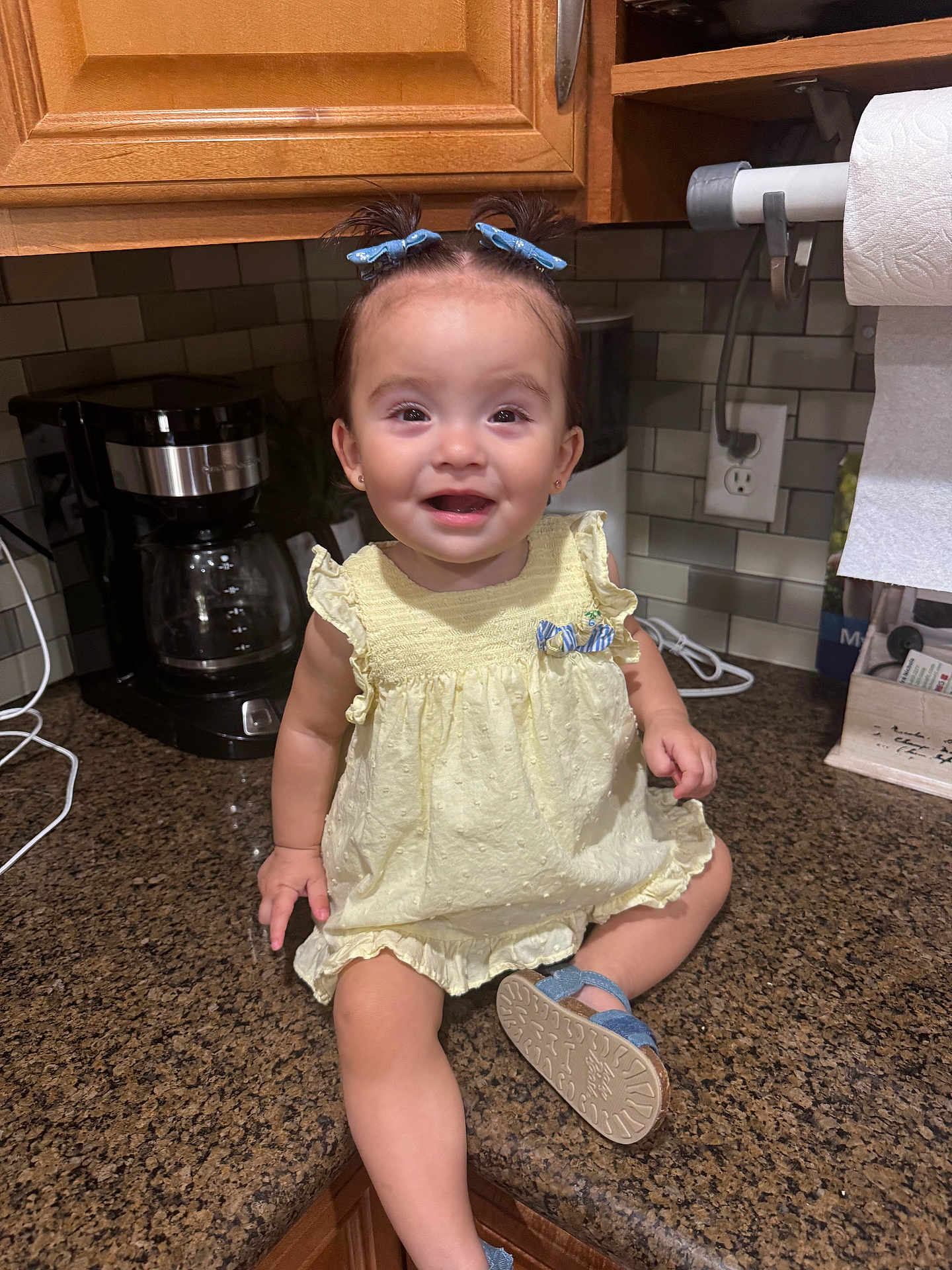 Gianna is registered to the contest to win money with this photo: toddler, child, baby, kitchen, countertop, yellow_dress, blue_bows, blue_sandals, smiling, sitting, coffee_maker, paper_towels, cabinet, tile_wall, cute, happy, indoor, person, hair_bows, playful