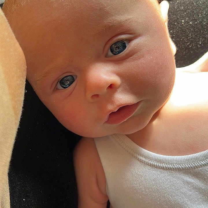 Saint is registered to the contest to win money with this photo: arm, baby, blue_eyes, casual, child, close_up, curious, cute, expression, face, human, indoor, infant, natural_light, newborn, portrait, skin, soft_skin, tattoo, white_clothing