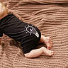 baby, blonde_hair, black_onesie, lightbulb_graphic, crawling, knitted_blanket, cozy, cute, infant, child, soft_texture, indoors, floor, barefoot, pattern, warm_colors, toddler, young_child, clothing, baby_pose