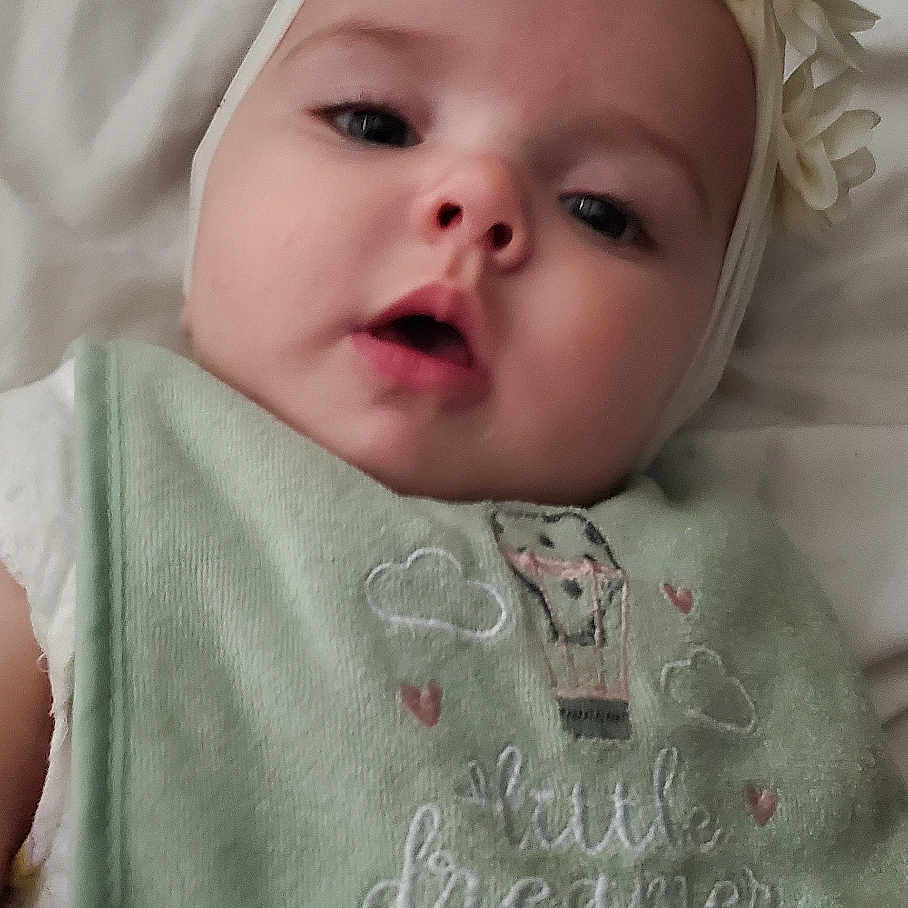 Mercee is registered to the contest to win money with this photo: baby, bib, blanket, child, clothing, cute, expression, face, flower, green, headband, indoors, infant, lying_down, portrait, sleepwear, soft, text, white, young