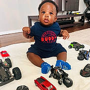 Jamal joined the competition — help win amazing prizes! baby, child, toy_car, toy_motorcycle, blue_socks, blanket, floor, living_room, television, furniture, curious, indoor, person, clothing, wooden_floor, sitting, cute, playtime, home, expression