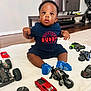 baby, child, toy_car, toy_motorcycle, blue_socks, blanket, floor, living_room, television, furniture, curious, indoor, person, clothing, wooden_floor, sitting, cute, playtime, home, expression