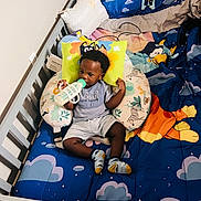Jamal is registered to the contest to win money with this photo: baby, bottle, bib, crib, child, clothing, colorful_bedding, cartoon_animals, cushion, face, footwear, furniture, indoors, infant, person, portrait, sleeping_area, socks, toy, young_child