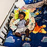 baby, bottle, bib, crib, child, clothing, colorful_bedding, cartoon_animals, cushion, face, footwear, furniture, indoors, infant, person, portrait, sleeping_area, socks, toy, young_child