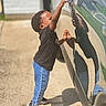 child, toddler, curious, car, car_handle, reflection, side_view, outdoor, sunny, jeans, black_shirt, sneakers, concrete, driveway, reflection_in_car, short_hair, standing, reaching, casual_clothing, daylight