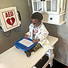 child, doctor, white_coat, stethoscope, glasses, magnetic_doodle_board, table, cabinet, aed_box, drawing, indoor, toy, playing, person, medical_theme, young_child, camouflage_pants, black_hair, footwear, focused