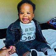 Jamal is registered to the contest to win money with this photo: baby, child, smiling, denim_jacket, denim_shorts, striped_socks, hand_holding, white_blanket, indoor, happy, tongue_out, person, cute, infant, portrait, clothing, sitting, bedroom, casual, joyful