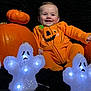 Elijah is registered to the contest to win money with this photo: baby, black_background, celebration, child, costume, cute, decor, face, festive, ghost_decoration, halloween, holiday, indoors, joy, light, orange_clothing, person, pumpkin, sitting, smiling