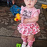 adult, baby, bow, carpet, casual, child, curious, expression, floor, footwear, girl, home, indoor, pink_clothing, plaything, room, seated_person, socks, standing, toy