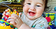 Quinn joined the competition — help win amazing prizes! baby, child, toy, colorful, smiling, happy, curly_hair, face, outdoor, play, seat, infant, cute, person, closeup, daylight, joyful, expression, clothing, background