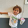 child, curly_hair, smiling, tshirt, shorts, drink, cup, straw, wall, texture, green_shoes, outdoor, happy, pose, person, casual_clothing, standing, cute, young, playful